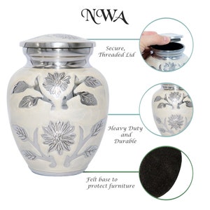 May include: White cremation urn with a silver floral design. The urn has a secure threaded lid and a felt base to protect furniture. "NWA" is printed on the top of the image.