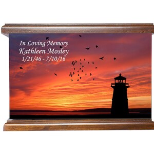May include: A wooden cremation urn featuring a sunset scene with a silhouette of a lighthouse. The urn is personalised with the inscription "In Loving Memory Kathleen Mosley 1/21/46-7/10/16".