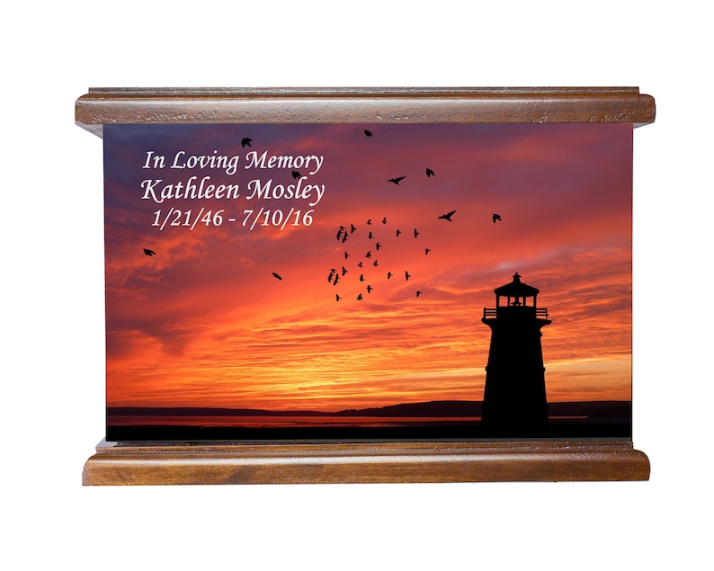 May include: A wooden cremation urn featuring a sunset scene with a silhouette of a lighthouse. The urn is personalised with the inscription "In Loving Memory Kathleen Mosley 1/21/46-7/10/16".