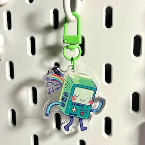 May include: A clear acrylic keychain featuring a green, smiling video game console character with blue arms and legs. The keychain has a green clasp and a design that includes a rainbow, musical notes, and the letters "BM".