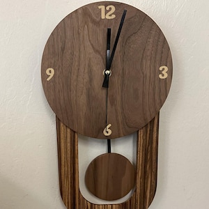 Può includere: Un orologio a pendolo in legno con un quadrante in noce e una cornice in legno scuro. Il quadrante dell'orologio ha numeri dorati e una lancetta delle ore e dei minuti nera. Il pendolo è in legno di noce.