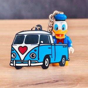 Könnte beinhalten: Ein Schlüsselanhänger mit einem blau-weißen Wohnwagen-Design mit einem roten Herz und einer Donald Duck-Cartoonfigur. Der Schlüsselanhänger hat eine silberne Kette und einen schwarzen Ring.