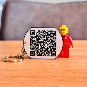 Könnte beinhalten: Ein weißer, kreisförmiger Schlüsselanhänger mit einem schwarzen QR-Code und einem silbernen Schlüsselring. Eine rote und gelbe Lego-Figur steht neben dem Schlüsselanhänger auf einer Holzoberfläche.