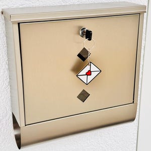 Könnte beinhalten: Ein metallisch-beiger Briefkasten mit Schlüssel und Schlüsselanhänger. Der Schlüsselanhänger zeigt ein weißes Umschlagdesign mit einem roten Herz. Der Briefkasten ist an einer weißen, strukturierten Wand befestigt.