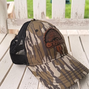 May include: A camouflage baseball cap with a black mesh back. The cap features a brown patch with a turkey graphic and the text "Only Fans". The hat is resting on a white wooden surface.