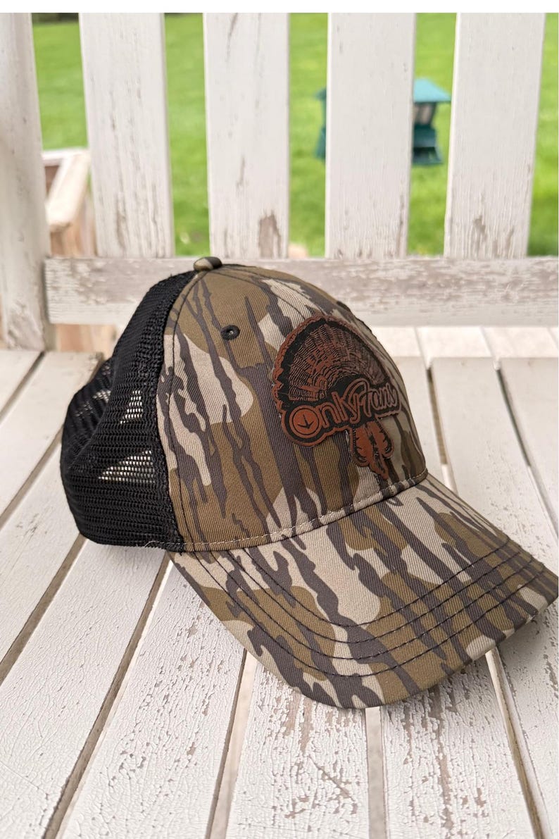 May include: A camouflage baseball cap with a black mesh back. The cap features a brown patch with a turkey graphic and the text "Only Fans". The hat is resting on a white wooden surface.