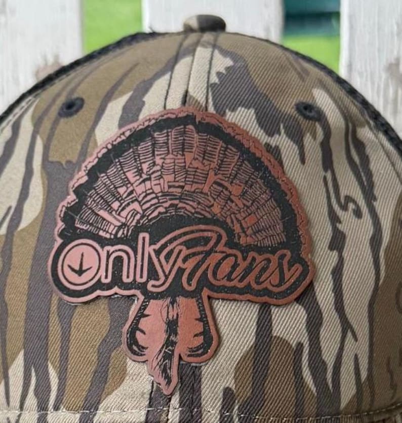 May include: Camouflage baseball cap with a brown and black patch featuring a turkey fan design and the text "OnlyFans". The cap has a curved brim and a mesh back.