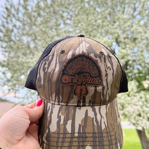 May include: Camouflage baseball cap with a black mesh back. The front of the cap features a brown leather patch with a turkey design and the text "Only Fans". The cap has a curved brim and is made of a camouflage pattern.