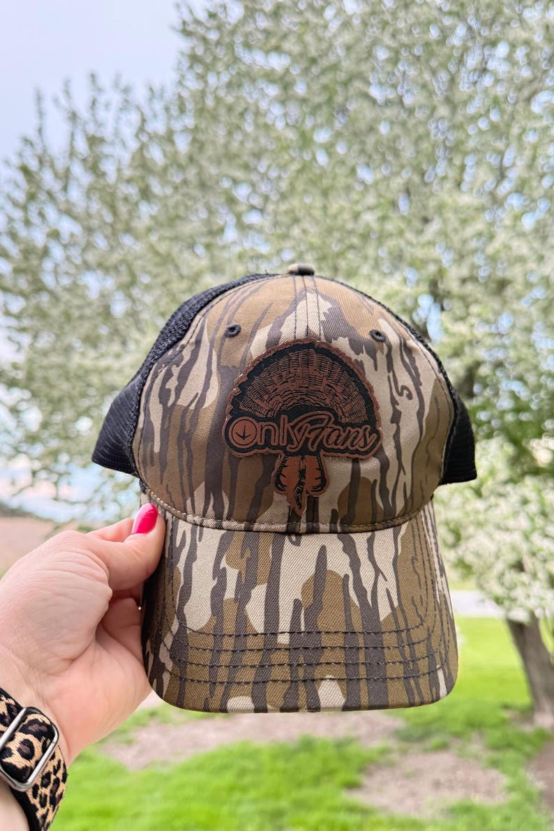May include: Camouflage baseball cap with a black mesh back. The front of the cap features a brown leather patch with a turkey design and the text "Only Fans". The cap has a curved brim and is made of a camouflage pattern.
