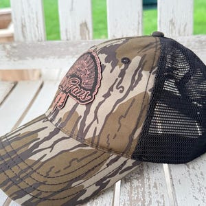 May include: A camouflage baseball cap with a black mesh back. The front of the cap features a patch with the words "Turkey Fans" in a cursive font, and a turkey graphic. The cap is resting on a white wooden surface.
