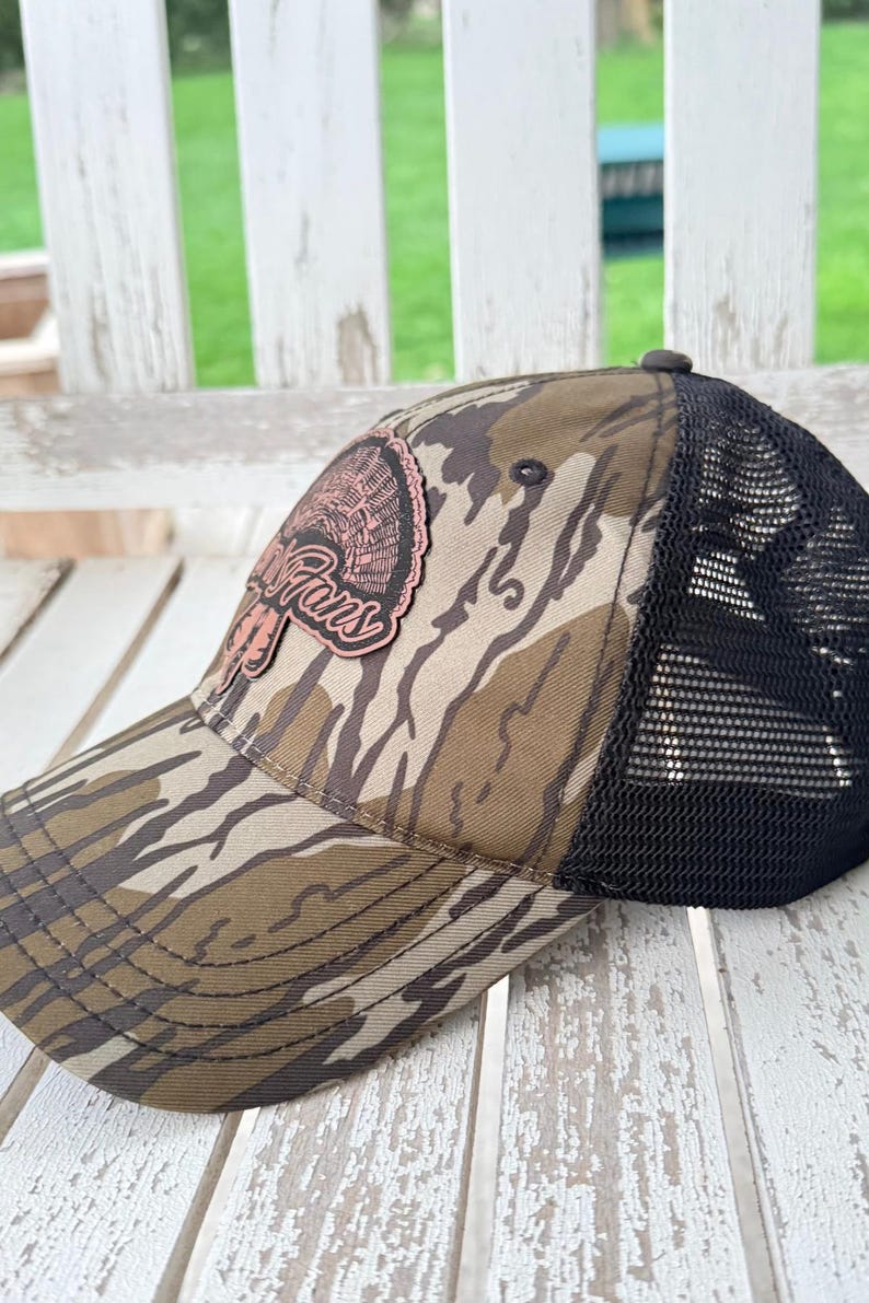 May include: A camouflage baseball cap with a black mesh back. The front of the cap features a patch with the words "Turkey Fans" in a cursive font, and a turkey graphic. The cap is resting on a white wooden surface.