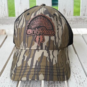 May include: A camouflage baseball cap with a black mesh back. The cap features a brown patch with a turkey illustration and the text "Only Fans" in a stylized font. The hat is designed with a curved brim.
