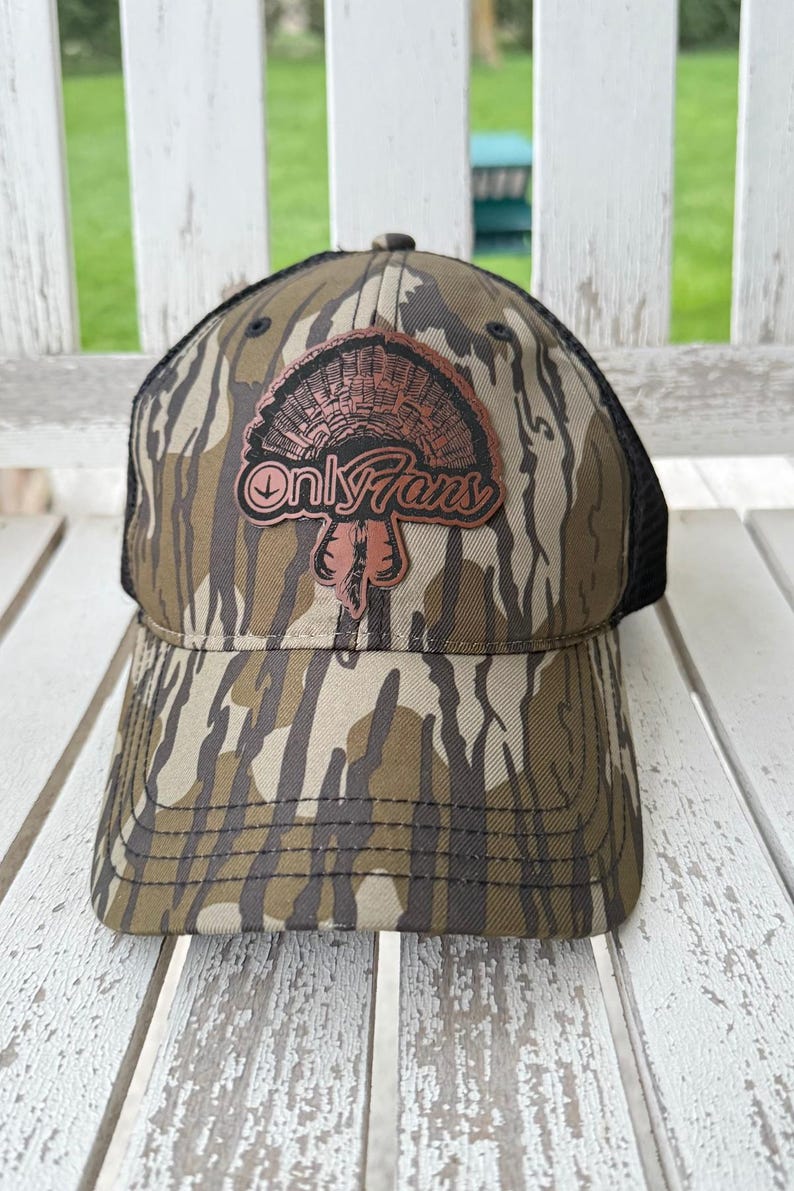 May include: A camouflage baseball cap with a black mesh back. The cap features a brown patch with a turkey illustration and the text "Only Fans" in a stylized font. The hat is designed with a curved brim.