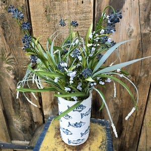 Op de afbeelding: Een cilindrische witte vaas met een getextureerd oppervlak voorzien van een herhalend patroon van donkerblauwe visillustraties. De vaas is gevuld met een arrangement van kunstmatig groen gras, witte takjes en trossen kleine donkerblauwe bloemen.