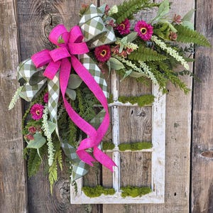 May include: A decorative wreath featuring a white-framed window design, adorned with vibrant pink flowers, green ferns, and a large pink bow. The window is set against a rustic wooden background, creating a charming, natural aesthetic.