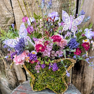 May include: A butterfly-shaped basket filled with a colourful floral arrangement. The flowers are in shades of pink, purple, and white, with a 'Happy Mother's Day' sign. The basket is lined with moss and decorated with lavender butterflies.
