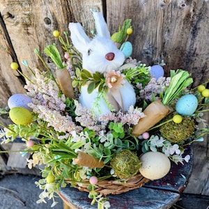 Könnte beinhalten: Ein dekorativer Osterkorb mit einem weißen Hasen, künstlichen Karotten und bunten Eiern. Der Korb ist mit Grün, rosa Blüten und gelben Akzenten gefüllt und schafft eine frühlingshafte Dekoration. Der Korb ist aus gewebtem Material.