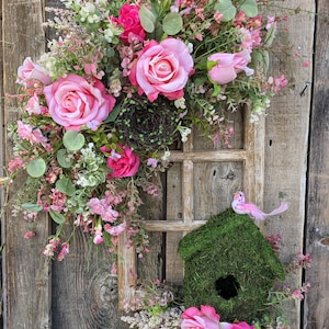 May include: A decorative floral wreath with pink roses, greenery, and a small birdhouse. The wreath is adorned with various pink flowers and greenery, creating a vibrant and natural aesthetic. The birdhouse is covered in green moss and has a small pink bird perched on top.