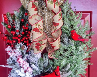 May include: A festive Christmas wreath with a mix of faux evergreen branches, red berries, and artificial snow. The wreath is adorned with a large bow featuring a cardinal bird pattern and two red cardinal figurines. The wreath is displayed on a red door.