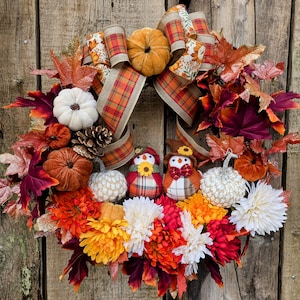 May include: A fall-themed wreath with a large plaid bow, pumpkins, and colorful leaves. The wreath features two owl figures, white and orange pumpkins, and a variety of fall-colored flowers. The background is a wooden surface.