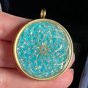 May include: A gold pendant with a turquoise blue geometric design inside. The pendant is round and has a loop at the top for attaching a chain.