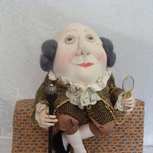 Puede incluir: Una mu&ntilde;eca hecha a mano con una cabeza grande, que lleva una chaqueta con estampado marr&oacute;n y dorado con un cuello de encaje blanco, sosteniendo un bast&oacute;n y una lupa. La mu&ntilde;eca est&aacute; sentada en un muro de ladrillo.