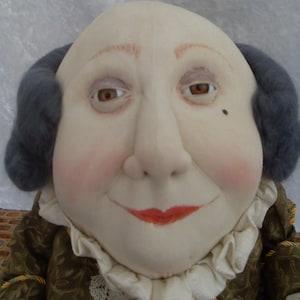 Puede incluir: Una mu&ntilde;eca con la cara redonda, peluca gris y un cuello de encaje blanco. La mu&ntilde;eca lleva una chaqueta de color marr&oacute;n y dorado con estampado.