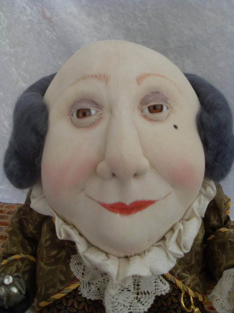 Puede incluir: Una mu&ntilde;eca con la cara redonda, peluca gris y un cuello de encaje blanco. La mu&ntilde;eca lleva una chaqueta de color marr&oacute;n y dorado con estampado.