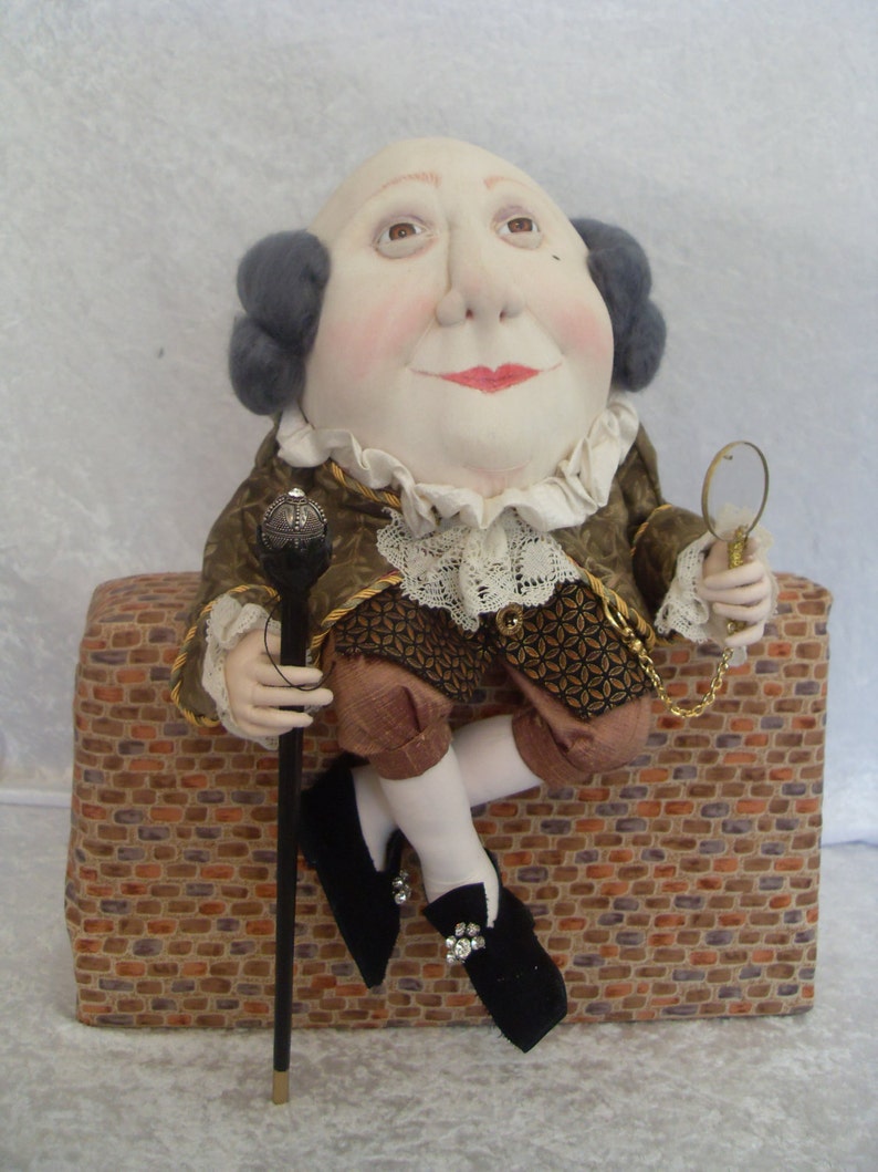 Puede incluir: Una mu&ntilde;eca hecha a mano con una cabeza grande, que lleva una chaqueta con estampado marr&oacute;n y dorado con un cuello de encaje blanco, sosteniendo un bast&oacute;n y una lupa. La mu&ntilde;eca est&aacute; sentada en un muro de ladrillo.