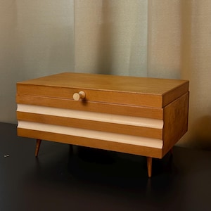 Könnte beinhalten: Eine Holzbox im Mid-Century-Stil mit hellbraunem Finish. Die Box hat eine rechteckige Form mit drei horizontalen cremefarbenen Streifen und einem kleinen Holzknopf. Sie steht auf vier dünnen, sich verjüngenden Beinen.