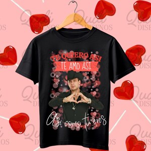 May include: Black t-shirt featuring a man making a heart shape with his hands. The shirt has the text "Te quiero asi, te amo asi, asi como tu eres" in white script. The background includes pink hearts and a pink background with heart-shaped lollipops.