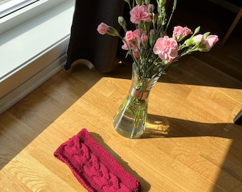 Könnte beinhalten: Ein weinrotes Stirnband mit Zopfmuster liegt auf einem Holzboden neben einer Glasvase mit rosa Nelken. Das strukturierte Design des Stirnbandes ist sichtbar, und die Blumen sorgen für einen Farbtupfer. Die Szene wird von natürlichem Licht beleuchtet.
