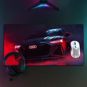 May include: A rectangular desk mat featuring a dark Audi car graphic with glowing red headlights and the Audi logo, accompanied by a black wired headset with a microphone and a white honeycomb-patterned gaming mouse placed on the surface.