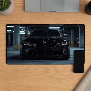 May include: A rectangular desk mat featuring a high-resolution print of a black BMW sports car parked in a dimly lit parking garage. The car is centered, showcasing its signature front grille, sharp LED headlights, and a sleek, glossy finish.