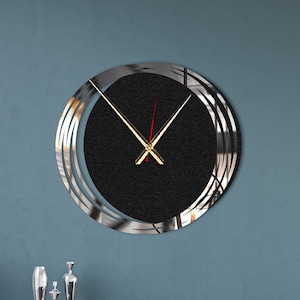 Op de afbeelding: Een moderne wandklok met een zwarte wijzerplaat en zilveren metalen ringen. De klok hangt aan een blauwe muur boven een donkerblauwe bank met kussens.