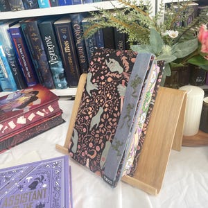K&ouml;nnte beinhalten: Ein h&ouml;lzerner Buchst&auml;nder h&auml;lt ein Buch mit einem schwarzen Einband mit einem Blumen- und Handdesign. Ein hellblaues Lesezeichen mit einem Baumdesign ist sichtbar. Der Hintergrund umfasst ein B&uuml;cherregal voller B&uuml;cher und dekorativer Blumen.