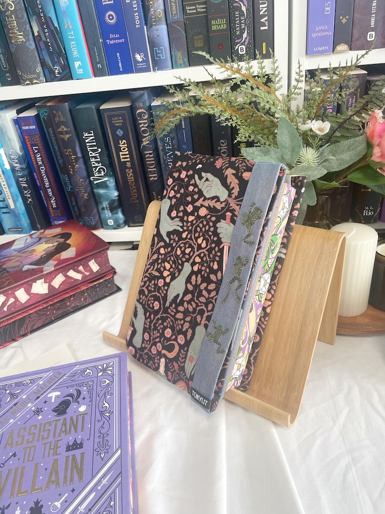 K&ouml;nnte beinhalten: Ein h&ouml;lzerner Buchst&auml;nder h&auml;lt ein Buch mit einem schwarzen Einband mit einem Blumen- und Handdesign. Ein hellblaues Lesezeichen mit einem Baumdesign ist sichtbar. Der Hintergrund umfasst ein B&uuml;cherregal voller B&uuml;cher und dekorativer Blumen.