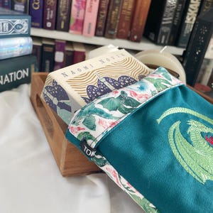 Peut inclure: Un sac à dos bleu sarcelle avec un imprimé floral blanc et vert et un dragon vert brodé sur le devant. Le sac est posé sur un plateau en bois avec un livre intitulé "Naomi Novik" sur le dessus du plateau.