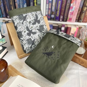 May include: Two book sleeves, one with a black and white crow pattern and the other with a green background and a black crow with a crescent moon above it.