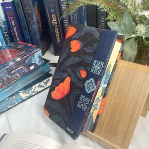 Puede incluir: Un libro con una cubierta oscura con amapolas naranjas y un marcapáginas azul con un diseño floral. El libro está apoyado en un soporte de madera, con otros libros visibles en el fondo. La palabra "TOKYLIT" está impresa en el libro.