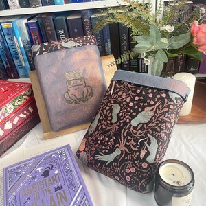 Könnte beinhalten: Zwei Buchhüllen, eine mit einem Frosch mit Krone und die andere mit einem Blumen- und Handdesign. Ein lila Buch mit dem Titel "Assistant to the Villain" ist sichtbar. Eine Kerze und Bücher sind im Hintergrund.