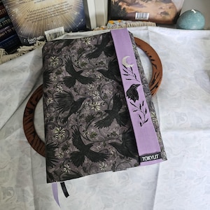 Può includere: Una copertina per libro in tessuto con un motivo grigio scuro di corvi neri, viticci e piccoli fiori bianchi. È presente una fascia elastica verticale color viola chiaro, ricamata con una mezzaluna argentata, rami frondosi e un corvo nero, con un'etichetta nera recante la scritta TOKYLIT.