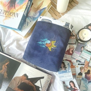 May include: A navy blue book sleeve with an embroidered sun and cloud design. The sleeve is surrounded by books, trading cards, and a lit candle. The books have titles and author names visible. The overall theme is fantasy and reading.