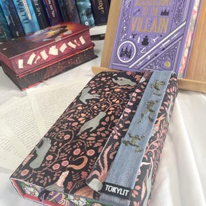 K&ouml;nnte beinhalten: Ein Buchcover mit einem floralen und Vogelmuster auf schwarzem Hintergrund. Ein Jeans-Lesezeichen mit goldenen Glitzerakzenten ist angebracht. Der Text "TOKYLIT" ist sichtbar. Weitere B&uuml;cher sind im Hintergrund.