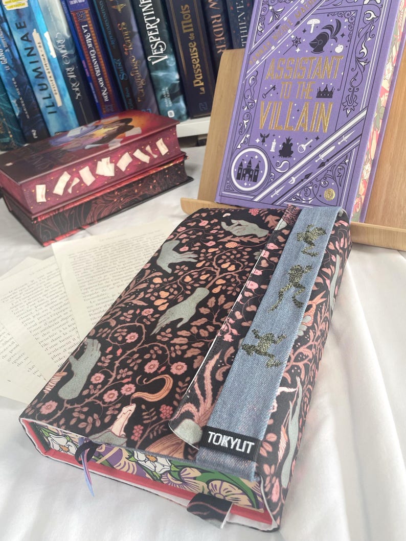 K&ouml;nnte beinhalten: Ein Buchcover mit einem floralen und Vogelmuster auf schwarzem Hintergrund. Ein Jeans-Lesezeichen mit goldenen Glitzerakzenten ist angebracht. Der Text "TOKYLIT" ist sichtbar. Weitere B&uuml;cher sind im Hintergrund.