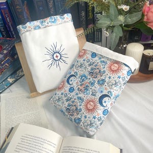 May include: Two celestial-themed book sleeves. One is white with a sunburst design, the other features a pattern of suns, moons, and clouds. Both have a white border. The sleeves are on a wooden stand, with an open book in the foreground.
