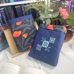 Puede incluir: Dos fundas de tela para libros. Una es azul marino con un estampado floral naranja, la otra es azul marino con un diseño floral bordado. Las fundas se exhiben frente a una estantería llena de libros.