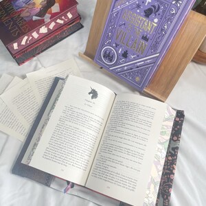 K&ouml;nnte beinhalten: Ein lila Hardcover-Buch mit dem Titel "Assistant to the Villain" in einem h&ouml;lzernen Buchst&auml;nder. Das Buch hat einen lila Einband mit goldfarbenen Buchstaben und dekorativen Elementen. Ein weiteres Buch ist ge&ouml;ffnet und zeigt bedruckte Seiten.