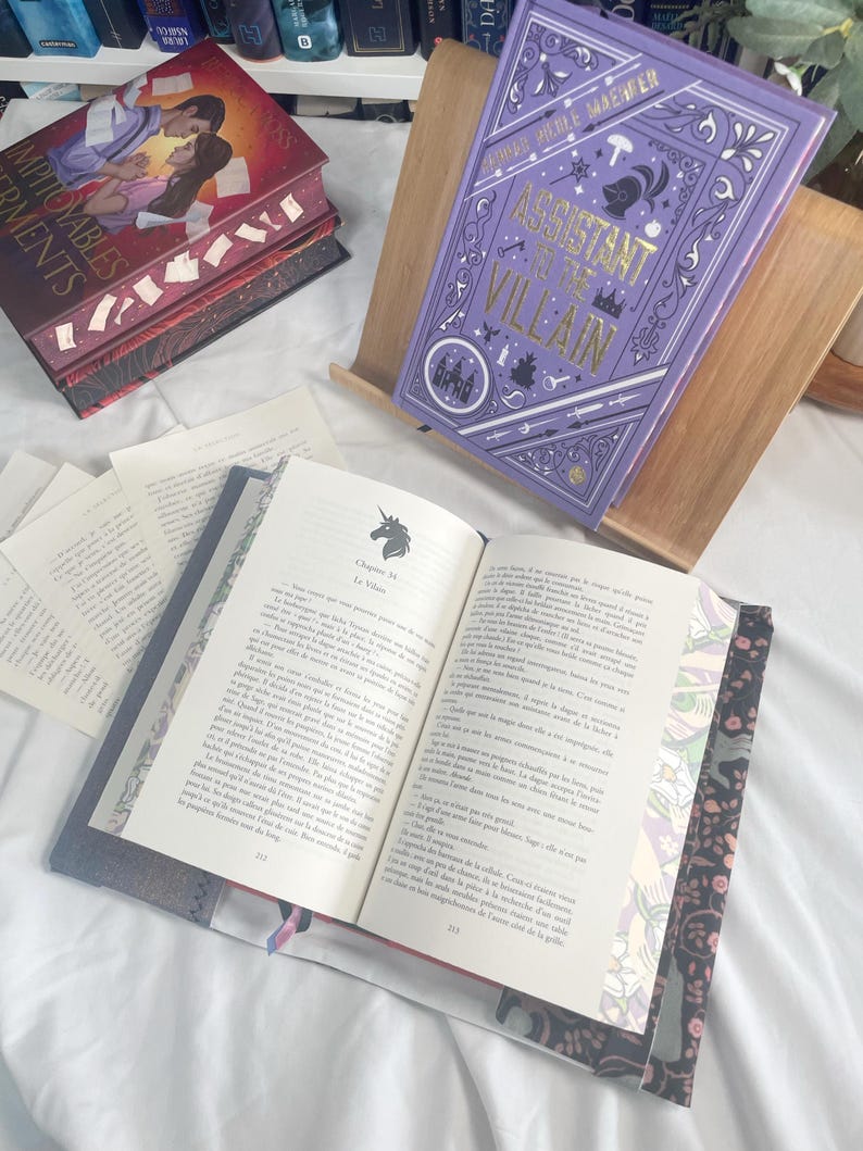K&ouml;nnte beinhalten: Ein lila Hardcover-Buch mit dem Titel "Assistant to the Villain" in einem h&ouml;lzernen Buchst&auml;nder. Das Buch hat einen lila Einband mit goldfarbenen Buchstaben und dekorativen Elementen. Ein weiteres Buch ist ge&ouml;ffnet und zeigt bedruckte Seiten.