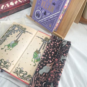 K&ouml;nnte beinhalten: Ein Hardcover-Buch mit dem Titel "Assistant to the Villain" in einem h&ouml;lzernen Buchst&auml;nder. Der Einband ist lila mit goldfarbenen Buchstaben und dekorativen Elementen. Offene Buchseiten zeigen Illustrationen von Fr&ouml;schen. Ein floraler Lesezeichen ist ebenfalls sichtbar.